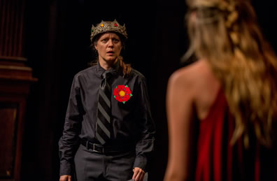 Henry in black shirt and pants with black and gray wide-striped tie and a red rose on his shirt and a crown on his head looks with concern on an unidentified woman, back to the camera.